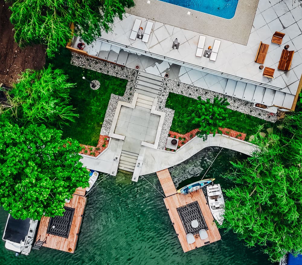 Aerial view of a modern lakeside patio with pool, steps, boats, and lush greenery.