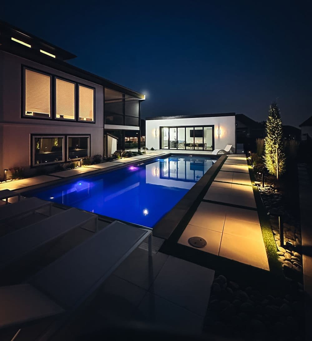 Modern home with a glowing blue pool at night, surrounded by stylish landscaping and lights.