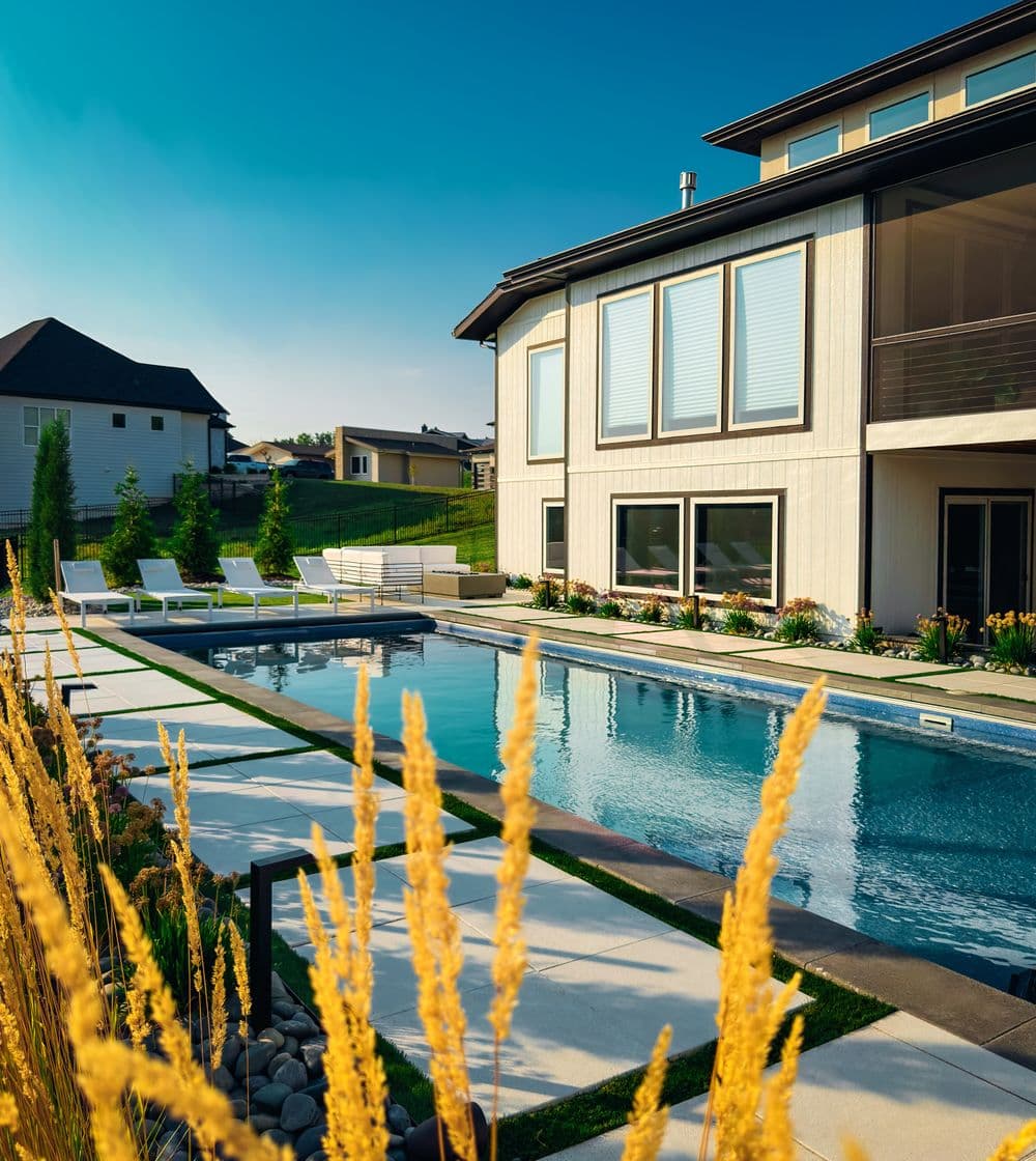 Modern home with a pool and landscaped yard, surrounded by greenery and sunlit skies.