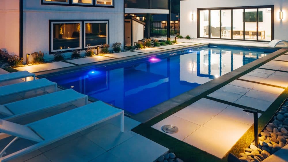 Modern backyard pool with lounge chairs, illuminated at night, surrounded by beautiful landscaping.