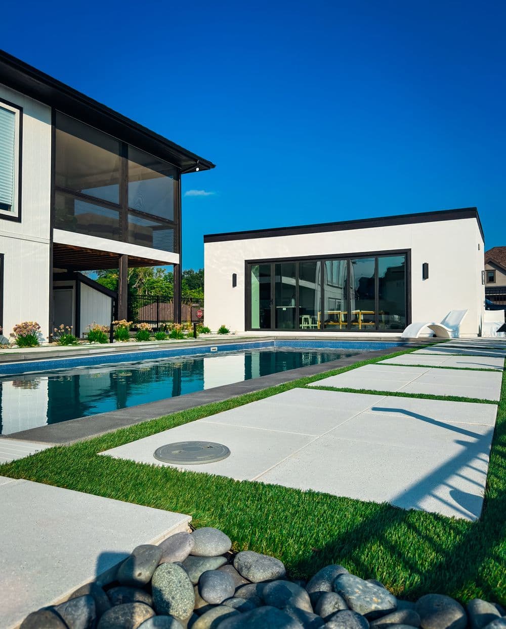 Modern backyard with pool, sleek white cottage, and lush green landscaping under a clear blue sky.