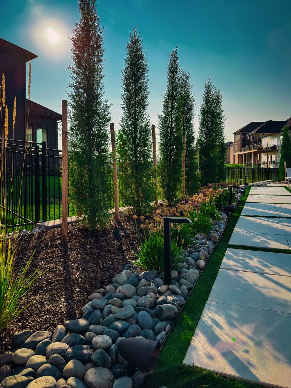 Lush garden with tall trees, landscaped stones, and modern architecture under a bright sky.