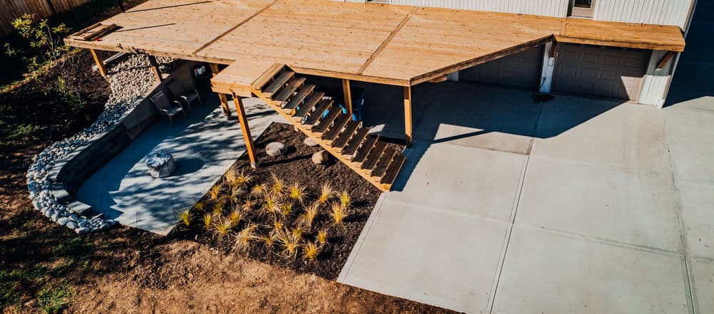 Aerial view of a wooden deck with stairs, landscaped area, and concrete driveway.