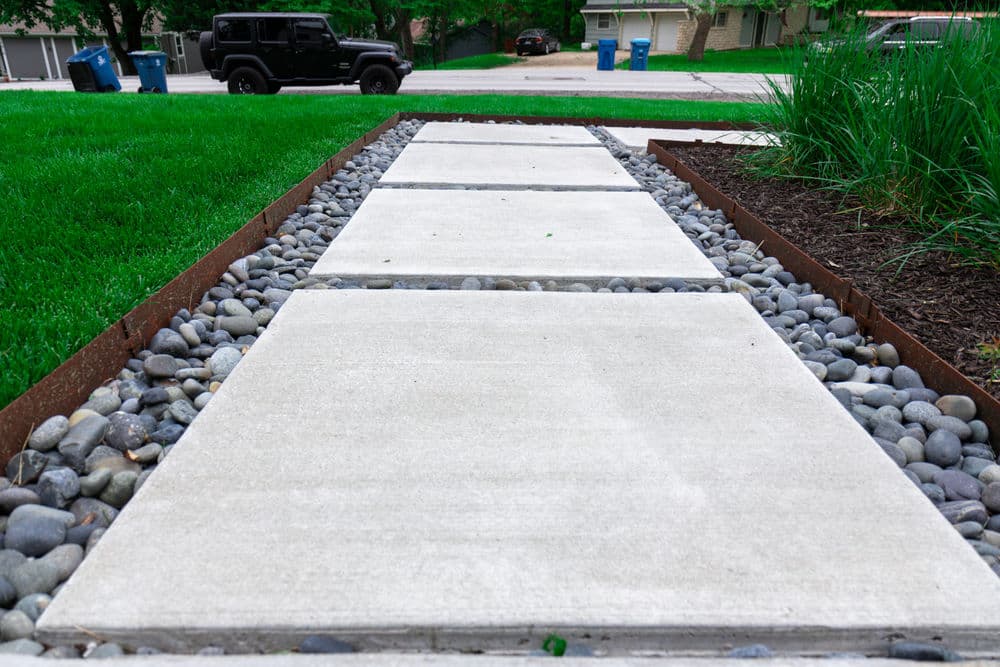 Concrete pathway lined with pebbles, surrounded by fresh grass and a parked Jeep.